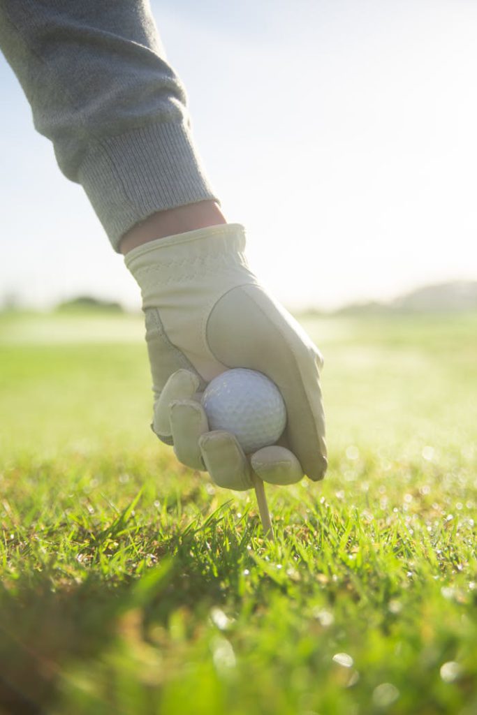 A gloved hand setting a golf ball on a tee in a sunny grassy field, perfect for sports enthusiasts.